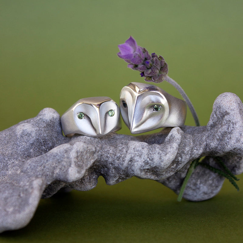 Handmade owl rings in sterling silver with sage green sapphires displayed on a stone with a lavender flower, part of the animal jewelry collection by Elina Gleizer.